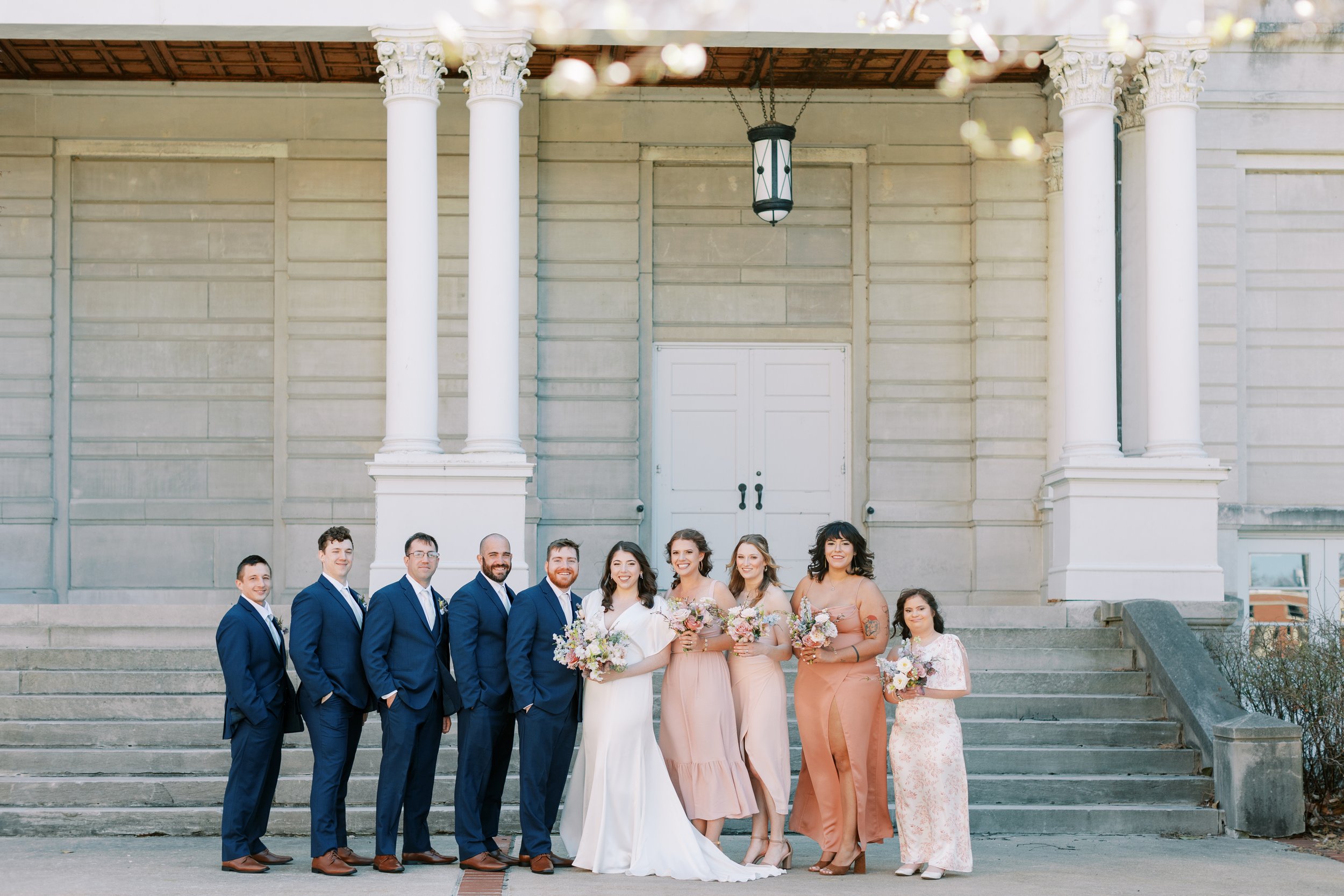 A bridal party is standing on the University of Missouri campus in Columbia, Missouri, posing for a photo on a wedding day. The bridal party is wearing navy and peach tones. Kiley Ann Photography captured the photo.