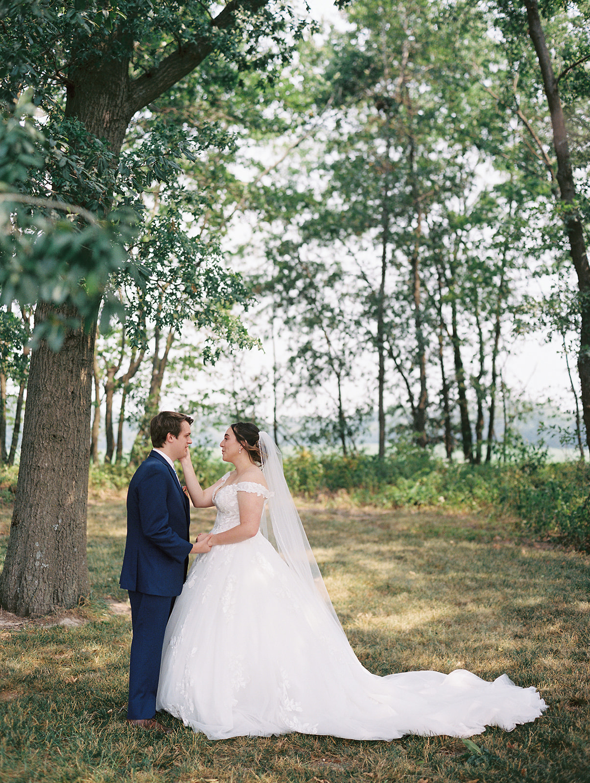 A bride and groom standing underneath trees on their wedding day at Emerson Fields, Missouri during their first look captured by Kiley Ann Photography. The groom is wearing a navy suit and the bride is wearing a princess ballgown.