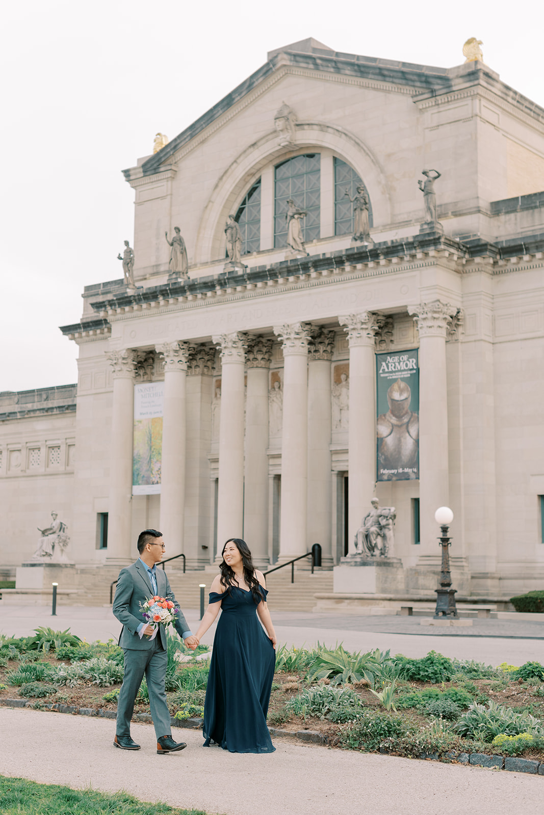 A newly engaged couple walking in front of the Art Museum in St. Louis posing for their engagement photos with Kiley Ann Photography. The couple is wearing a grey suit and a navy blue dress.