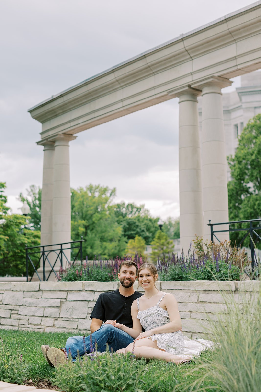 A couple sitting on a blanket in grass posing for their engagement session in Jefferson City Missouri with the Missouri Capitol in the background