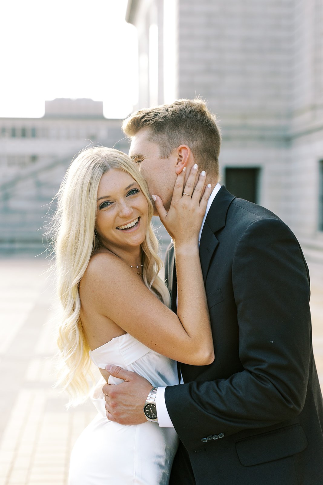 A bride to be laughing as her fiancé whispers something funny in her ear during their engagement session in Downtown, St. Louis