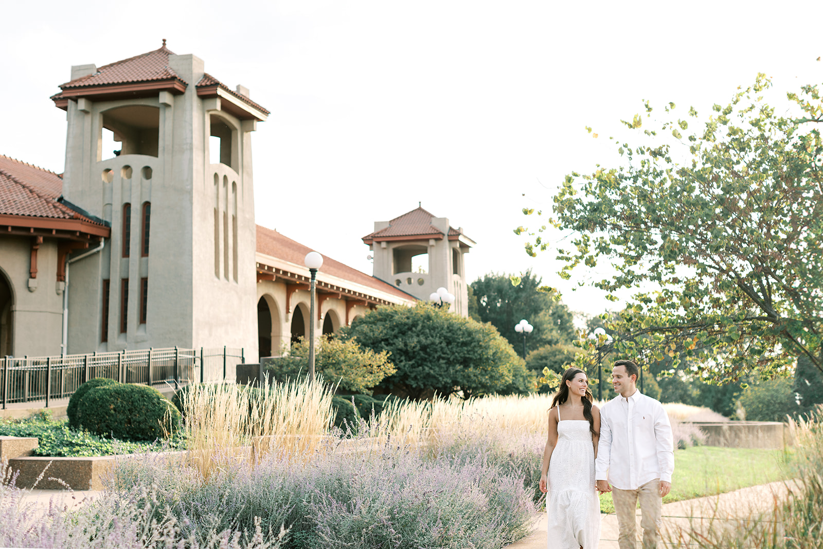 A couple walking during their engagement session at the World's Fair Pavilion in St. Louis, Missouri. The couple's attire consists of a white dress, a white button-up shirt, and khakis. This summer engagement session was captured by Kiley Ann Photography.